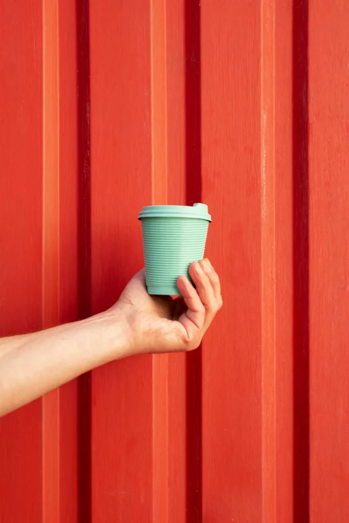 Mano sosteniendo una taza de café reutilizable de color verde menta contra una pared de metal acanalado de color rojo vibrante