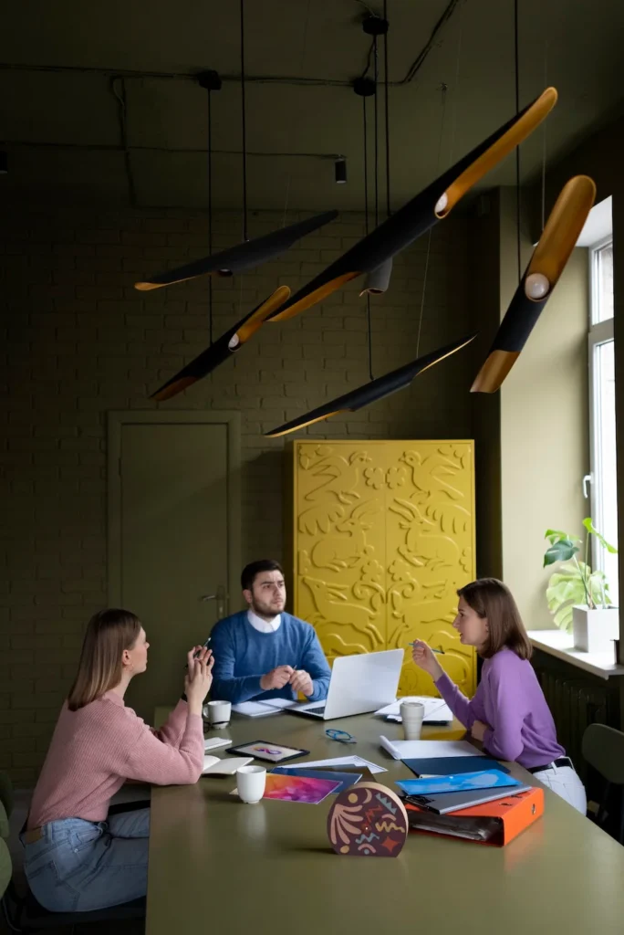 Tres profesionales de marketing en una reunión de equipo creativa en una oficina moderna. Discuten estrategias de marca alrededor de una mesa de conferencias con un portátil, carpetas de colores y material impreso. El fondo presenta una pared de ladrillo verde oliva y un armario amarillo tallado con luces colgantes contemporáneas.